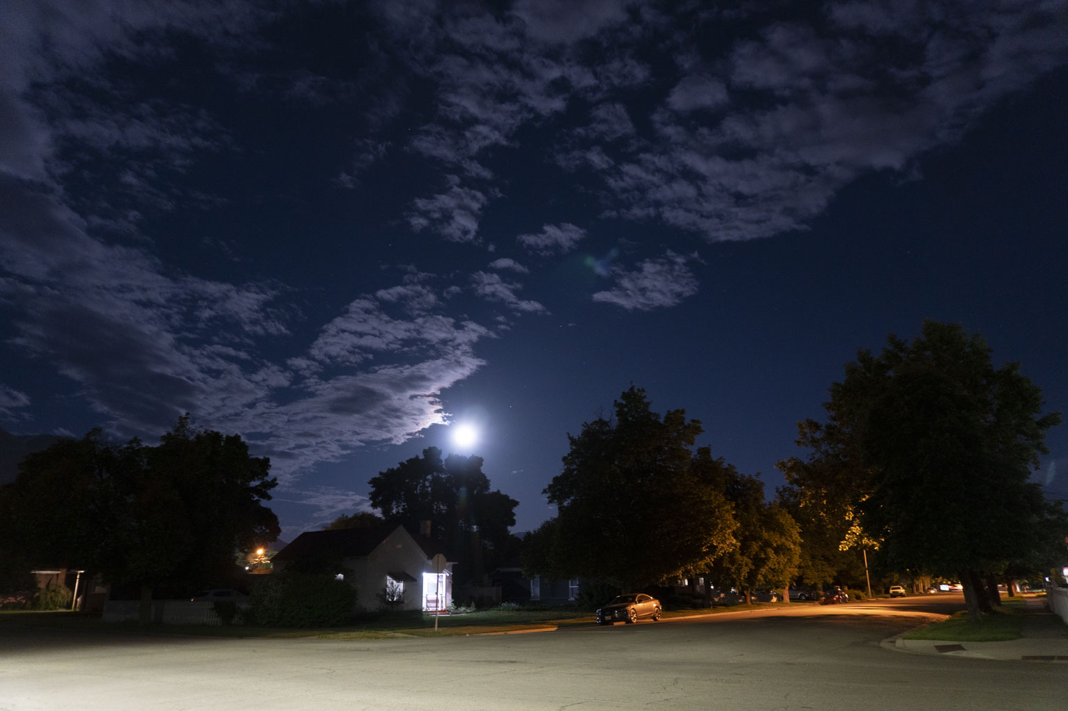 The crossroads of a neighborhood at night, the moon, low over houses, lights the clouds